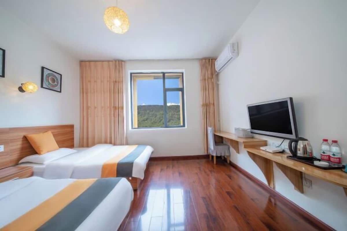 A hotel room with two beds, a television, a wooden desk, and a window with a view of greenery.