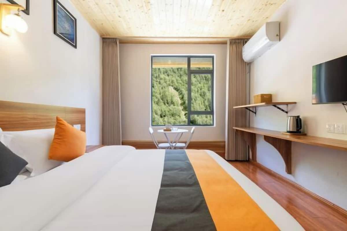A hotel room with a wooden ceiling, a large window with a forest view, a bed with white bedding, an orange pillow, a small table, and a TV on the wall.