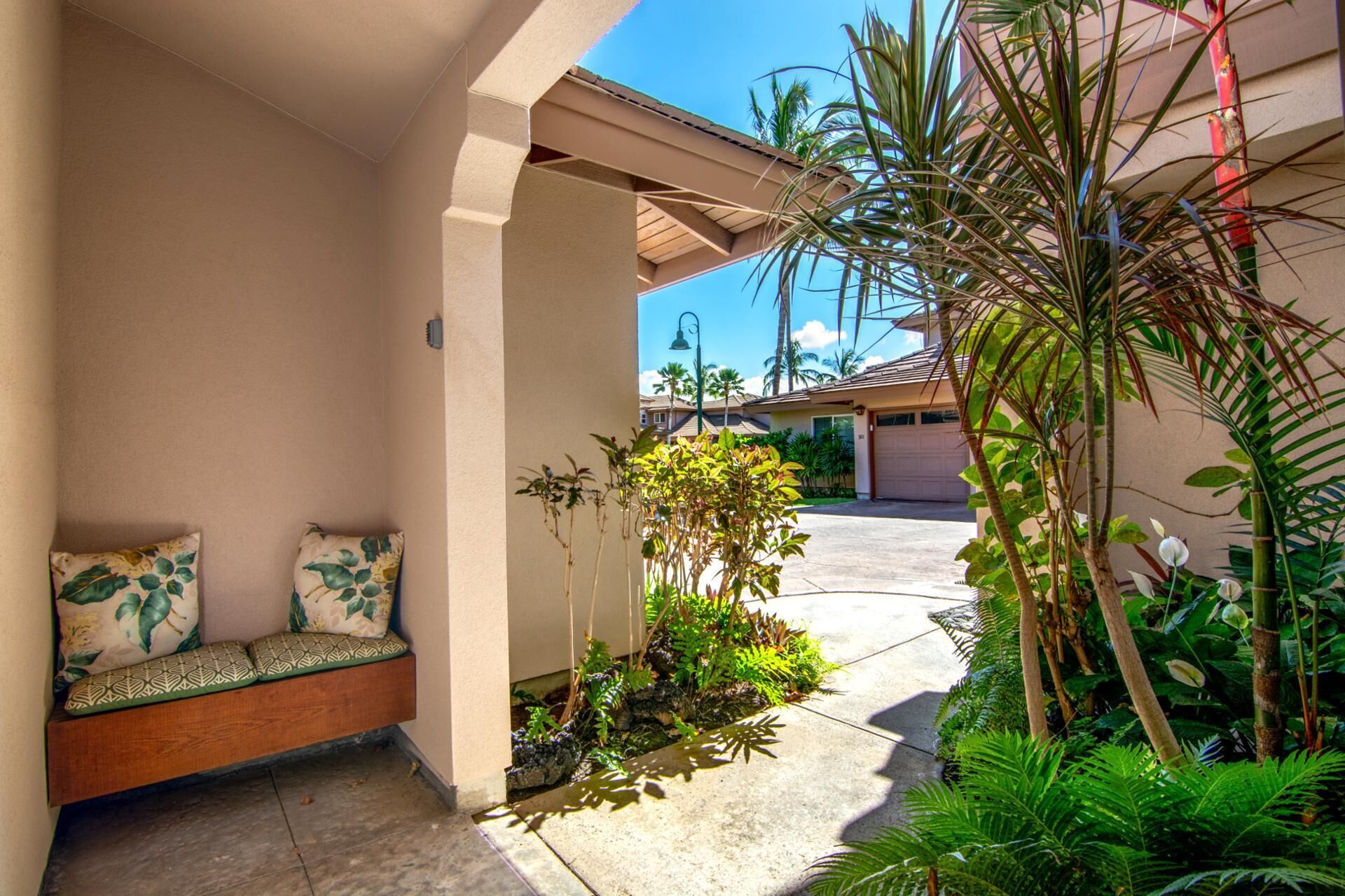 Un espacio al aire libre cubierto con un banco y cojines, rodeado de plantas tropicales y vistas despejadas a una calle residencial.