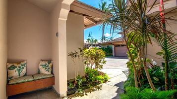 Un espacio al aire libre cubierto con un banco y cojines, rodeado de plantas tropicales y vistas despejadas a una calle residencial.