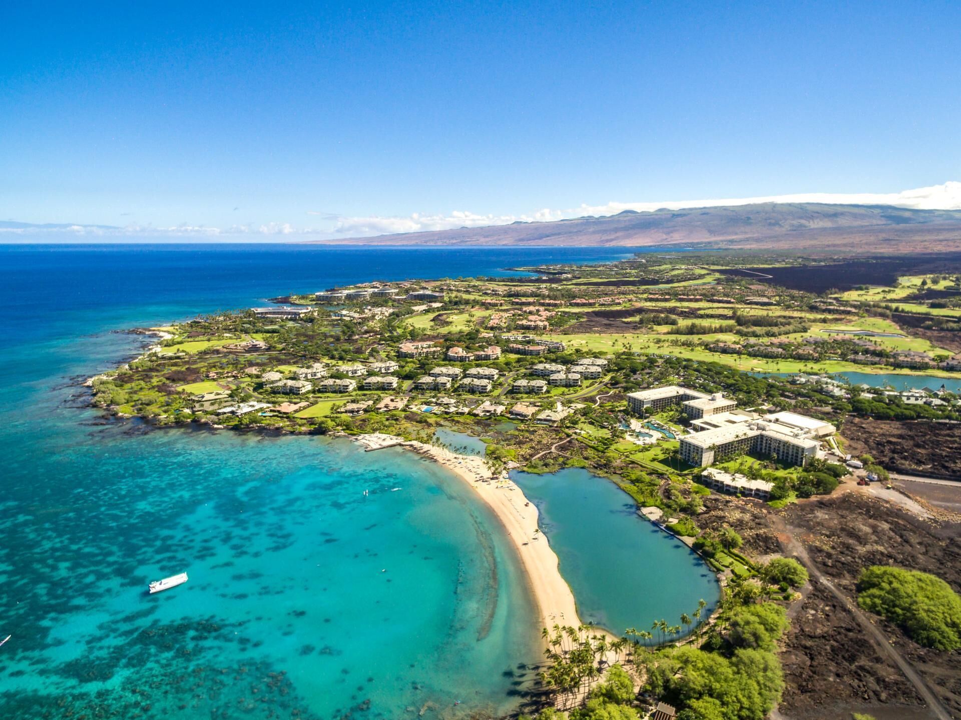 An aerial view of a coastal resort with a beach, clear turquoise waters, and a mix of residential and commercial buildings.