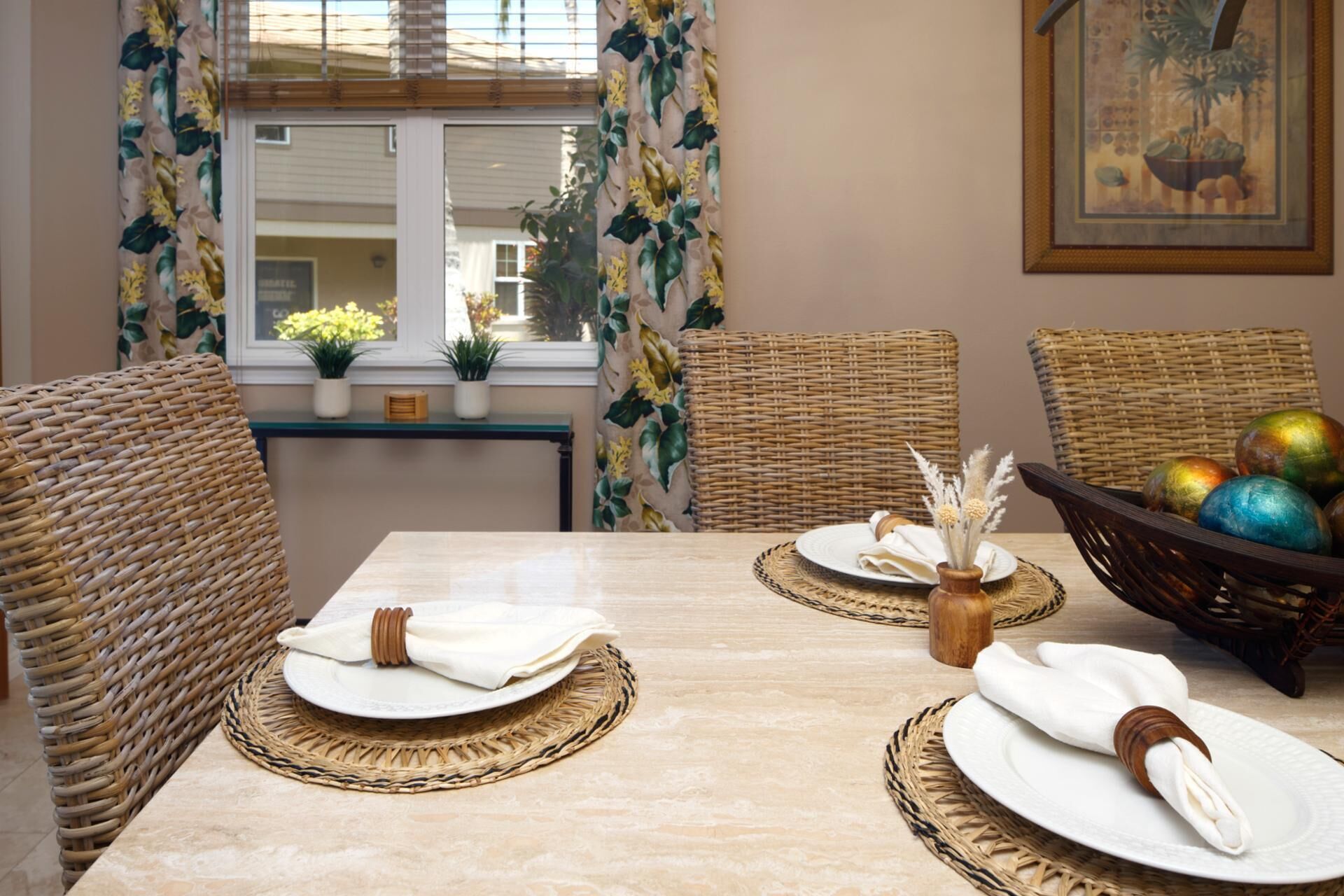 Un comedor con una mesa dispuesta para cuatro personas, rodeada de sillas de mimbre, una ventana con cortinas florales y un cuadro en la pared.