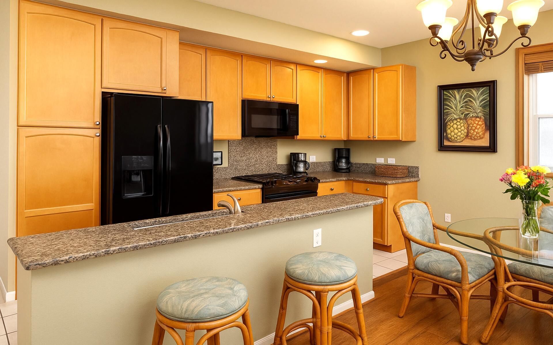 A kitchen with wooden cabinets, a black refrigerator, a microwave, a stove, and a dining area with wicker chairs and a glass table.