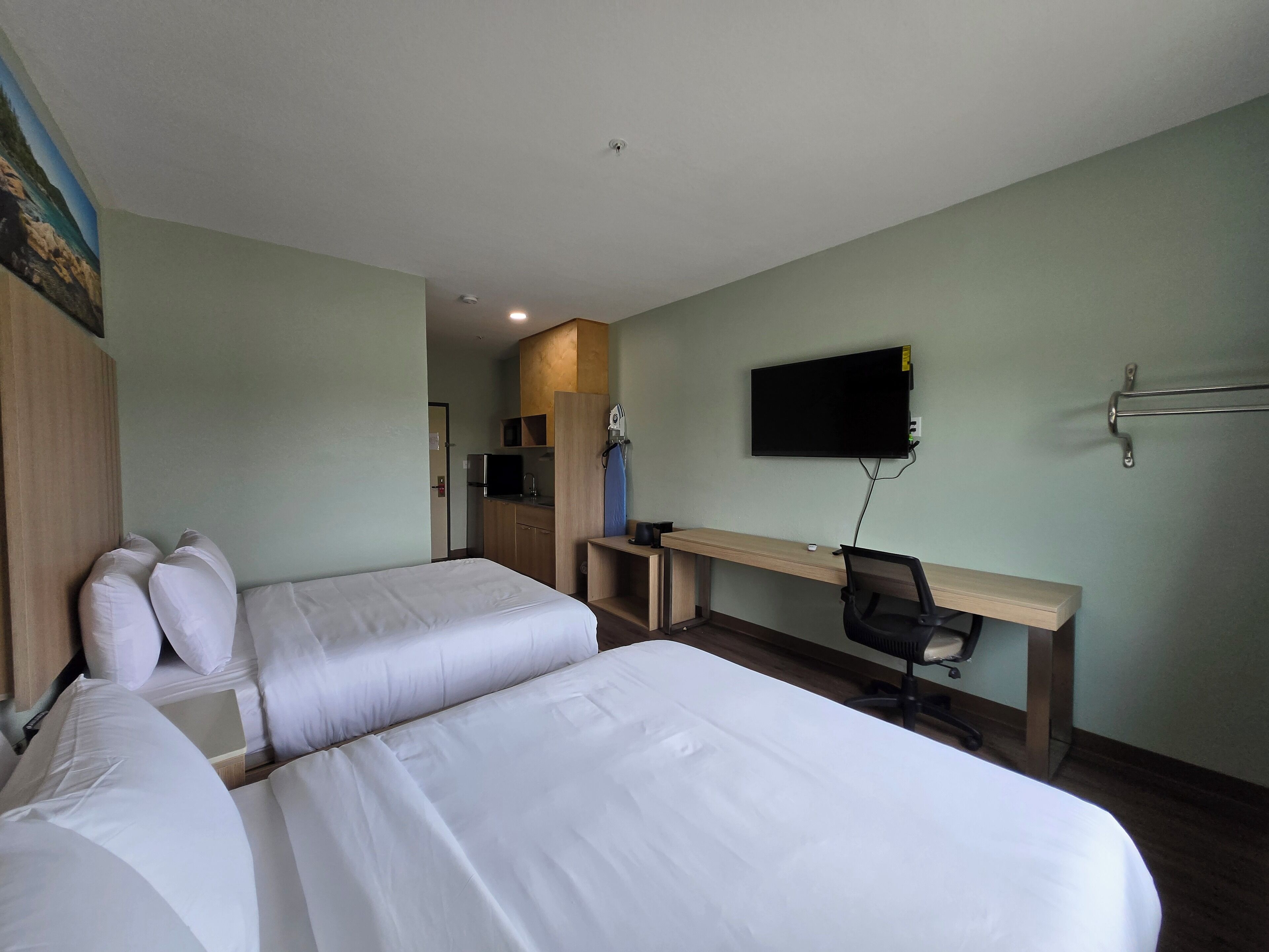 A hotel room with two beds, a desk, a chair, a TV, and a kitchenette.