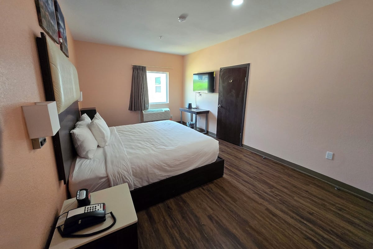 A hotel room with a bed, bedside table with a phone, a desk with a chair, a TV, and a window with curtains.