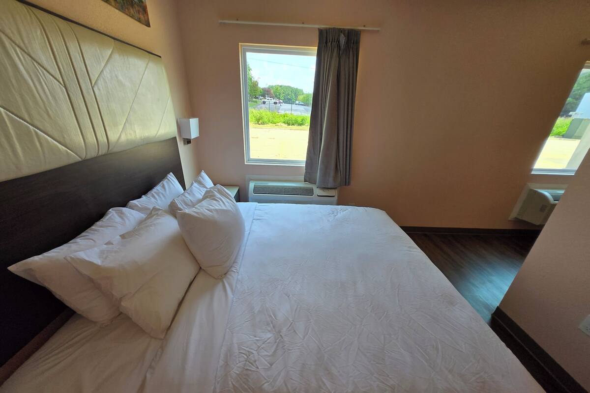 A hotel room with a large bed, a headboard, a window with curtains, and an air conditioning unit.