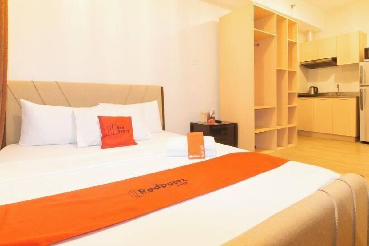 A hotel room with a bed, a nightstand, a red throw pillow, and a kitchen area with cabinets and a sink.