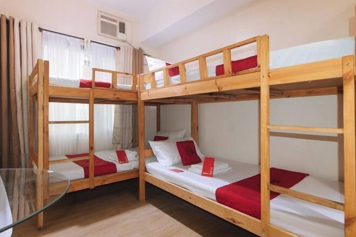 A bunk bed room with two beds, each with a red blanket and white pillows. There is a window with curtains, a radiator, and a glass table.