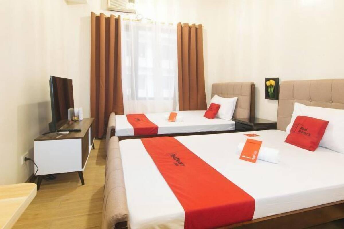 A hotel room with two beds, each with a red bedspread and matching pillows. There is a wooden nightstand with a lamp and a small table with a book and a phone. The room has a window with brown curtains and a picture of yellow flowers on the wall.