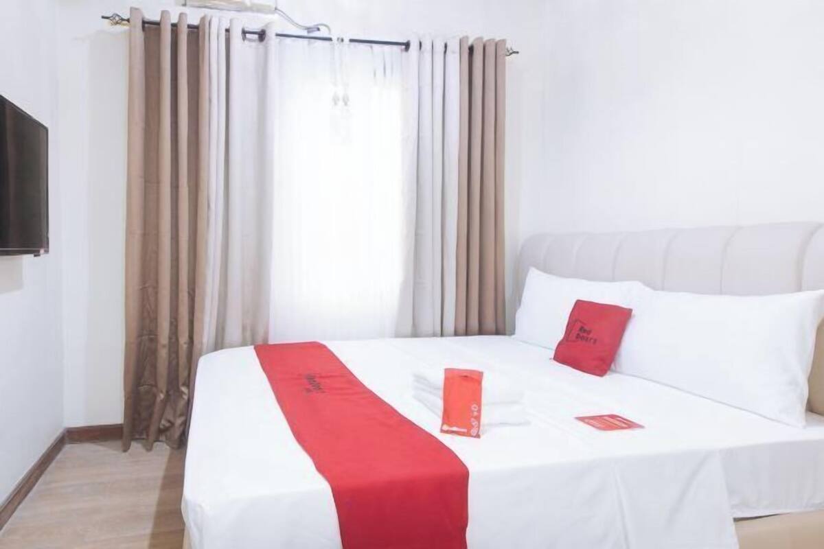 A hotel room with a bed, white bedding, a red throw, a red pillow, and a window with curtains.