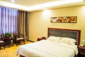 A hotel room with a large bed, a nightstand, a chair, a small table with a plant, and a painting on the wall. - Bali Hotel (Cao County)