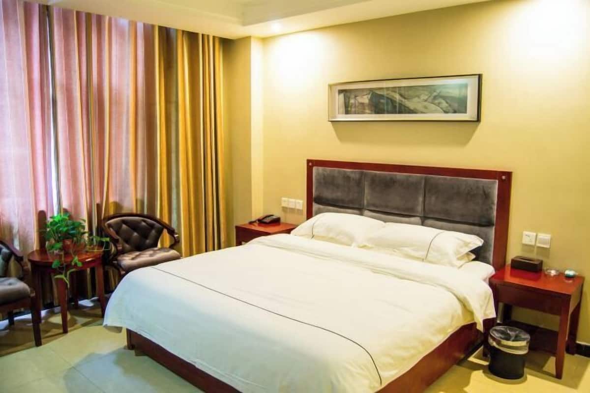 Une chambre d’hôtel avec un grand lit, une chaise, une petite table avec une plante, une table de chevet et une poubelle.