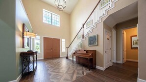Ein geräumiges Foyer mit einer monumentalen Treppe, einem Klavier und einer großen Holztür.