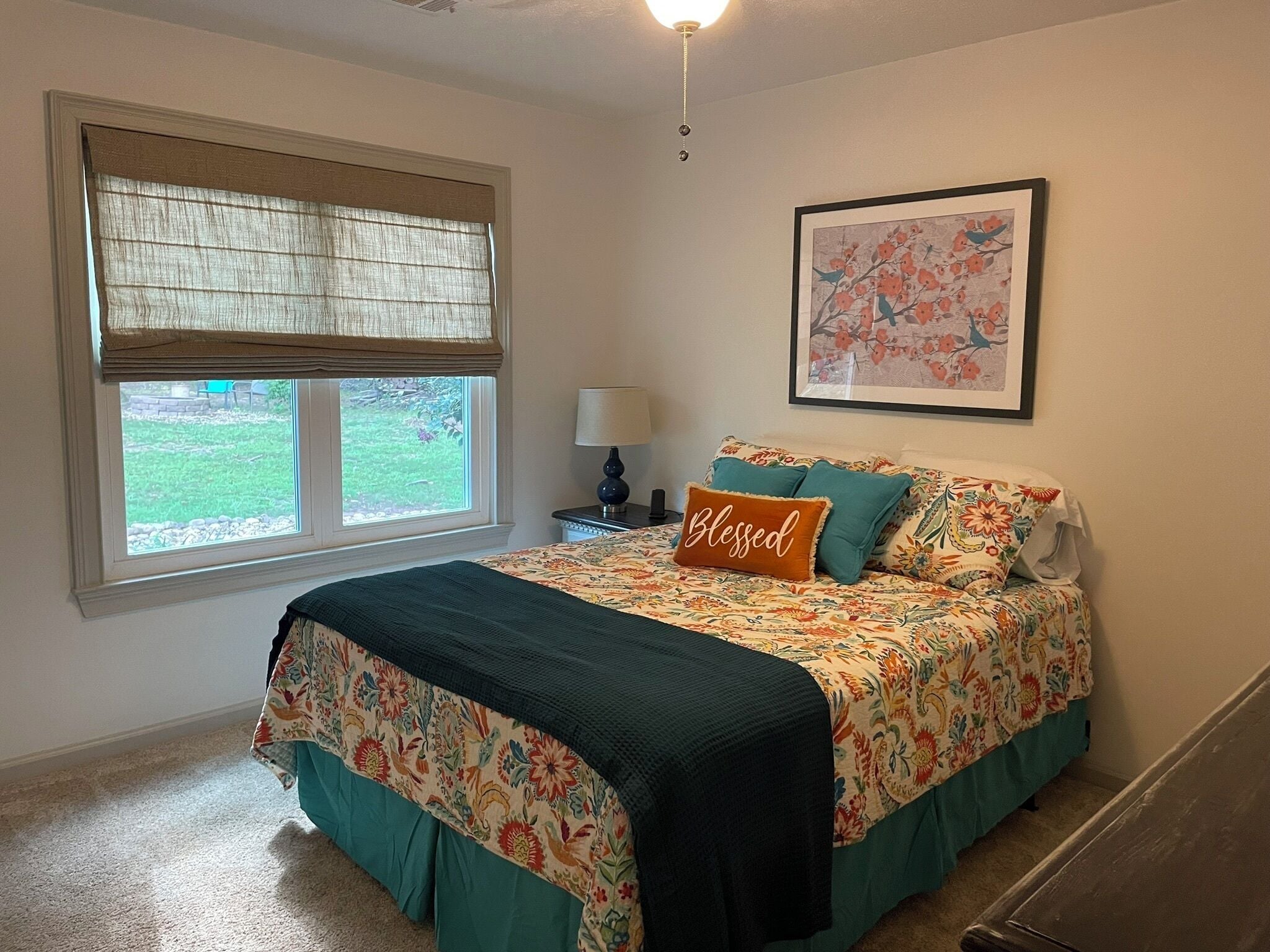 A bedroom with a bed, a window with blinds, a bedside table with a lamp, and a framed picture on the wall.
