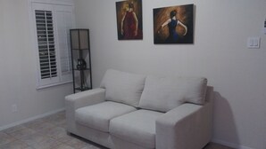 Een woonkamer met een beige bank, een vloerlamp en twee schilderijen aan de muur.