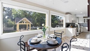 Un coin repas avec une table ronde dressée pour quatre personnes, de grandes fenêtres et une vue sur une maison et des arbres.