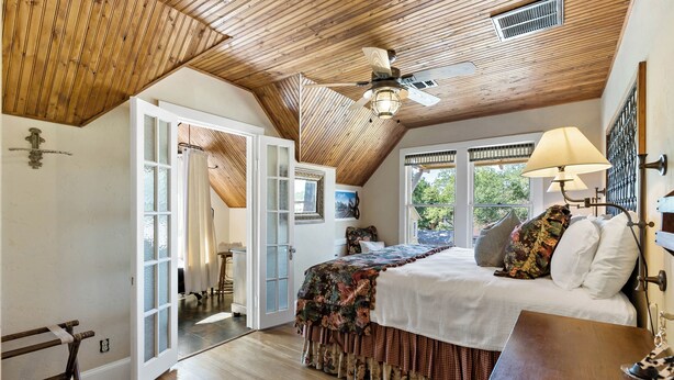 Une chambre avec un plafond en bois, un lit recouvert d’une literie à motifs floraux, une table de chevet et une fenêtre donnant sur un paysage verdoyant.