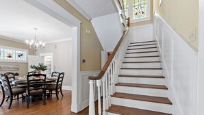Un escalier avec des balustrades blanches et des marches en bois, menant à un palier agrémenté d’un lustre et à un coin repas avec une table ronde et des chaises.
