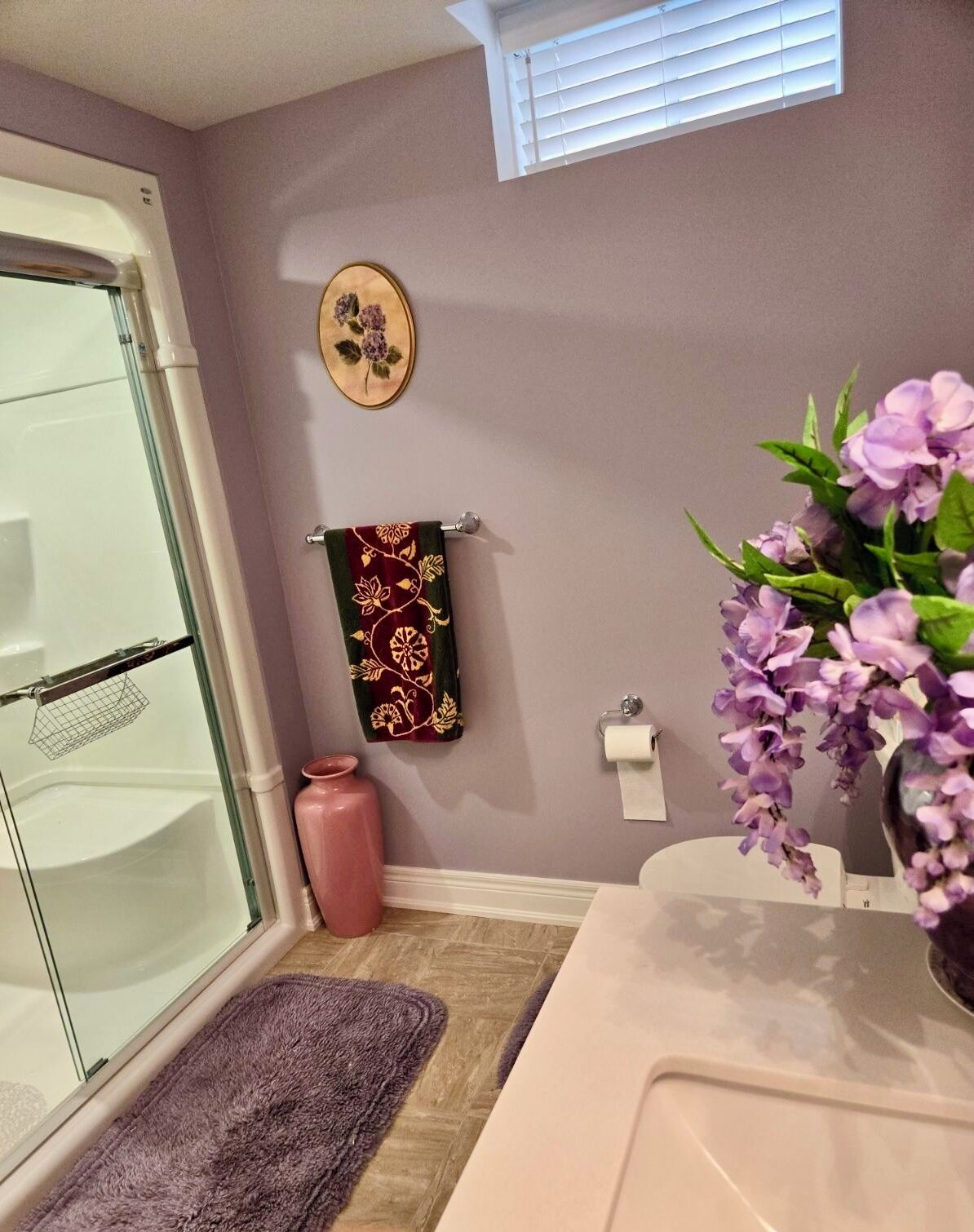 Une salle de bain avec une porte de douche en verre, un vase rose contenant des fleurs violettes, une serviette à motifs floraux et une petite plante en pot.