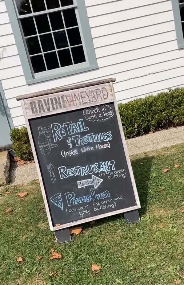 En skylt för vingården Ravine Vineyard, med information om försäljning, vinprovningar, restaurang och pizzaugn, samt pilar som visar vägen.