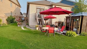 A residential backyard with a patio area featuring a table and chairs, a red umbrella, and a wooden deck. - Charming B&B with WiFi/Kitchenette and AC in Marvelous Niagara-on-the-Lake (Niagara-on-the-Lake)