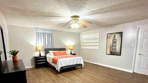 Un dormitorio con cama, mesitas de noche, ventilador de techo, ventana con cortinas y una puerta.