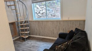 Un salon avec un canapé gris foncé, un coussin décoratif à motifs, un plancher en bois, une fenêtre donnant sur des rochers et des arbres, et un escalier en colimaçon.