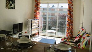 A living room with a dining table set, a TV, a window with curtains, and a colorful chair. - Limoges,, T2 ++parkings au Jardin ,,acces Facile ! (Limoges)