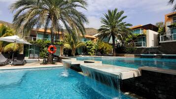 A swimming pool with water features, palm trees, and modern buildings in the background.
