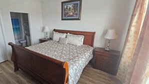 Um quarto com uma cama de madeira, duas mesas de cabeceira, um criado-mudo, uma porta e um quadro na parede.