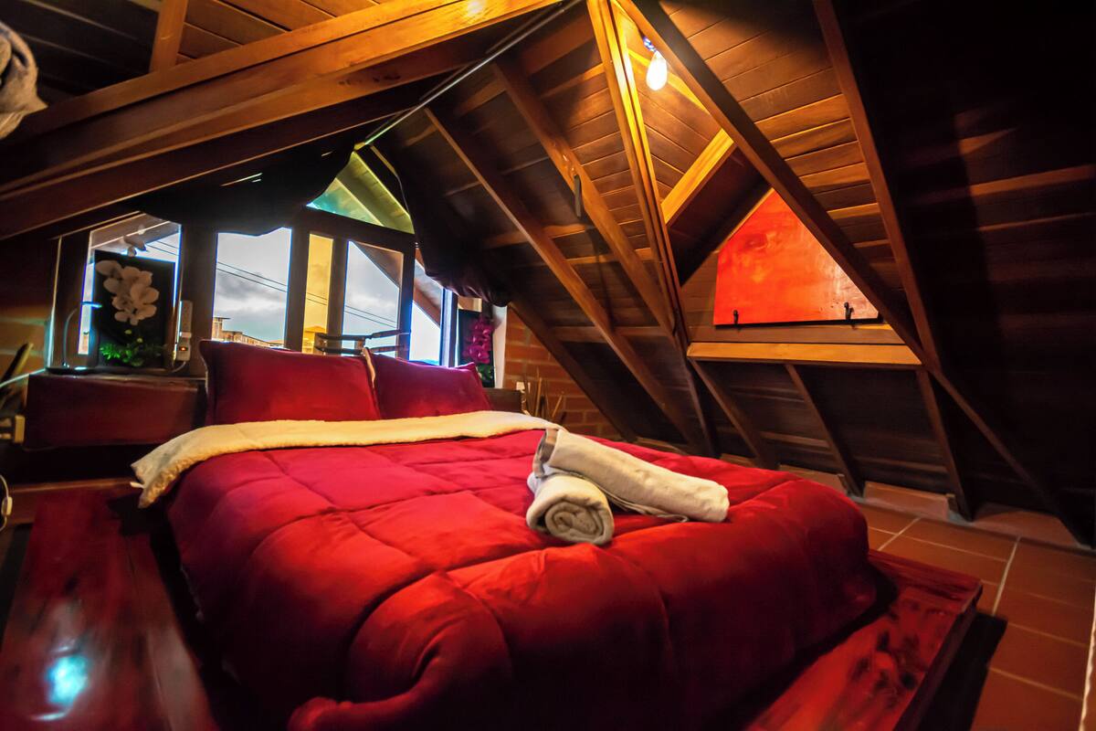 Quarto com colcha vermelha, vigas de madeira e claraboia.