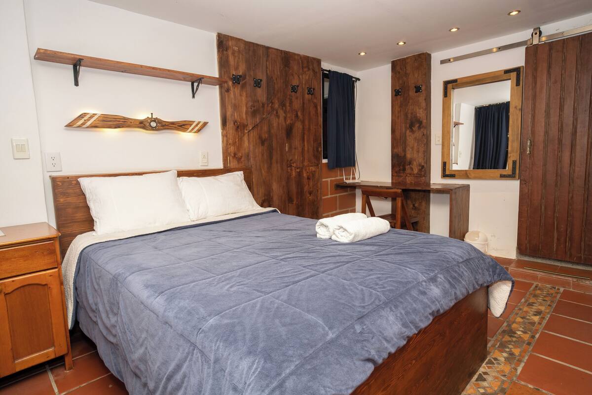 Quarto com cama de madeira, mesa de cabeceira, penteadeira, espelho e luminária fixada na parede.