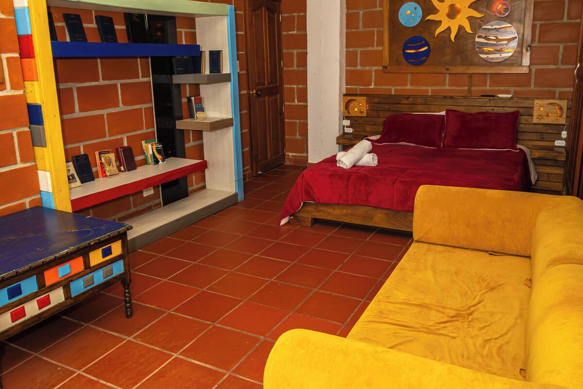 Um quarto com parede de tijolos, cama de madeira, sofá amarelo, mesa pequena e prateleira com livros.