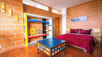 Una habitación con pared de ladrillo, una cama con manta roja, una mesa colorida y un estante con juguetes.