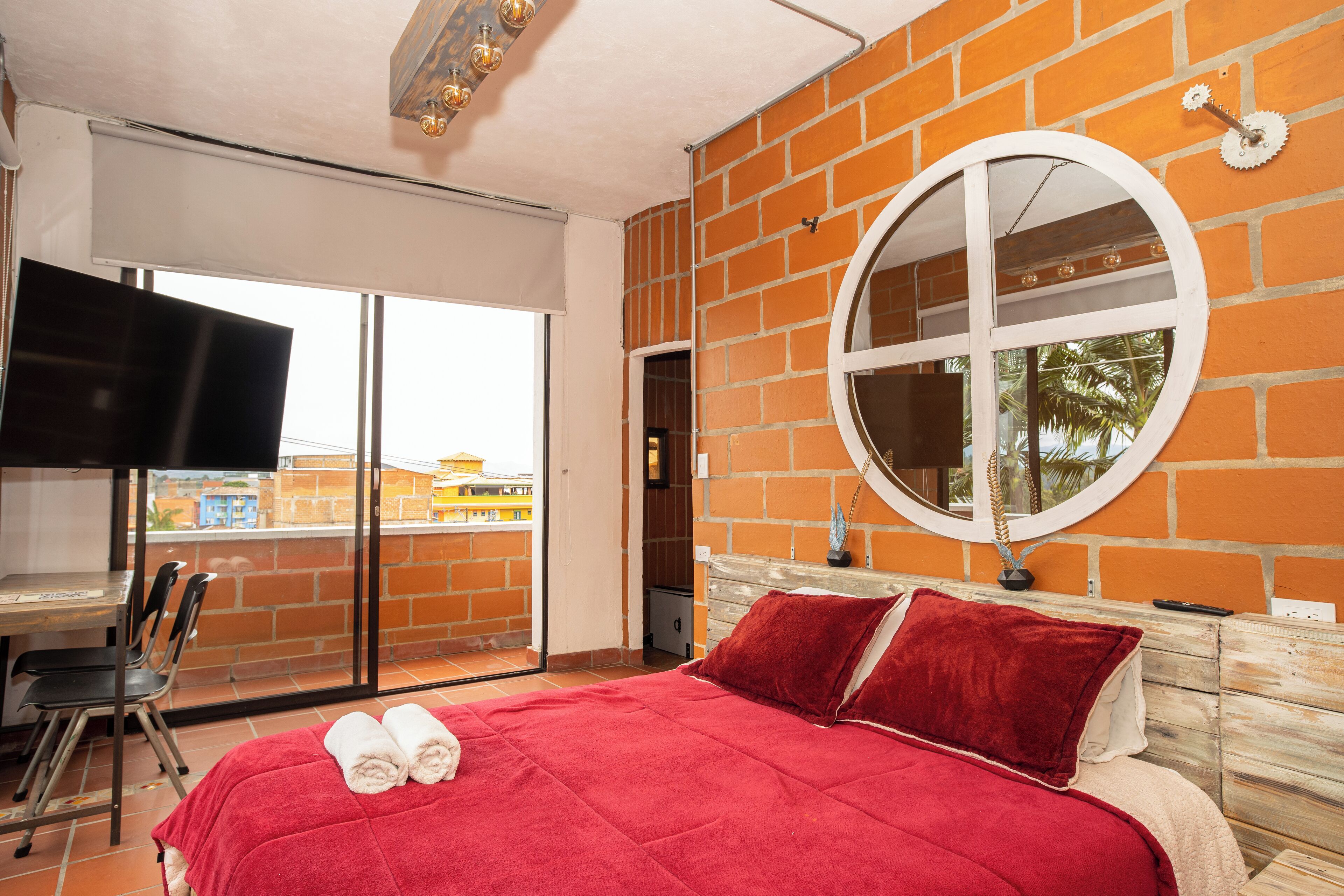 Un dormitorio con colcha roja, espejo redondo, televisor y balcón con vistas.