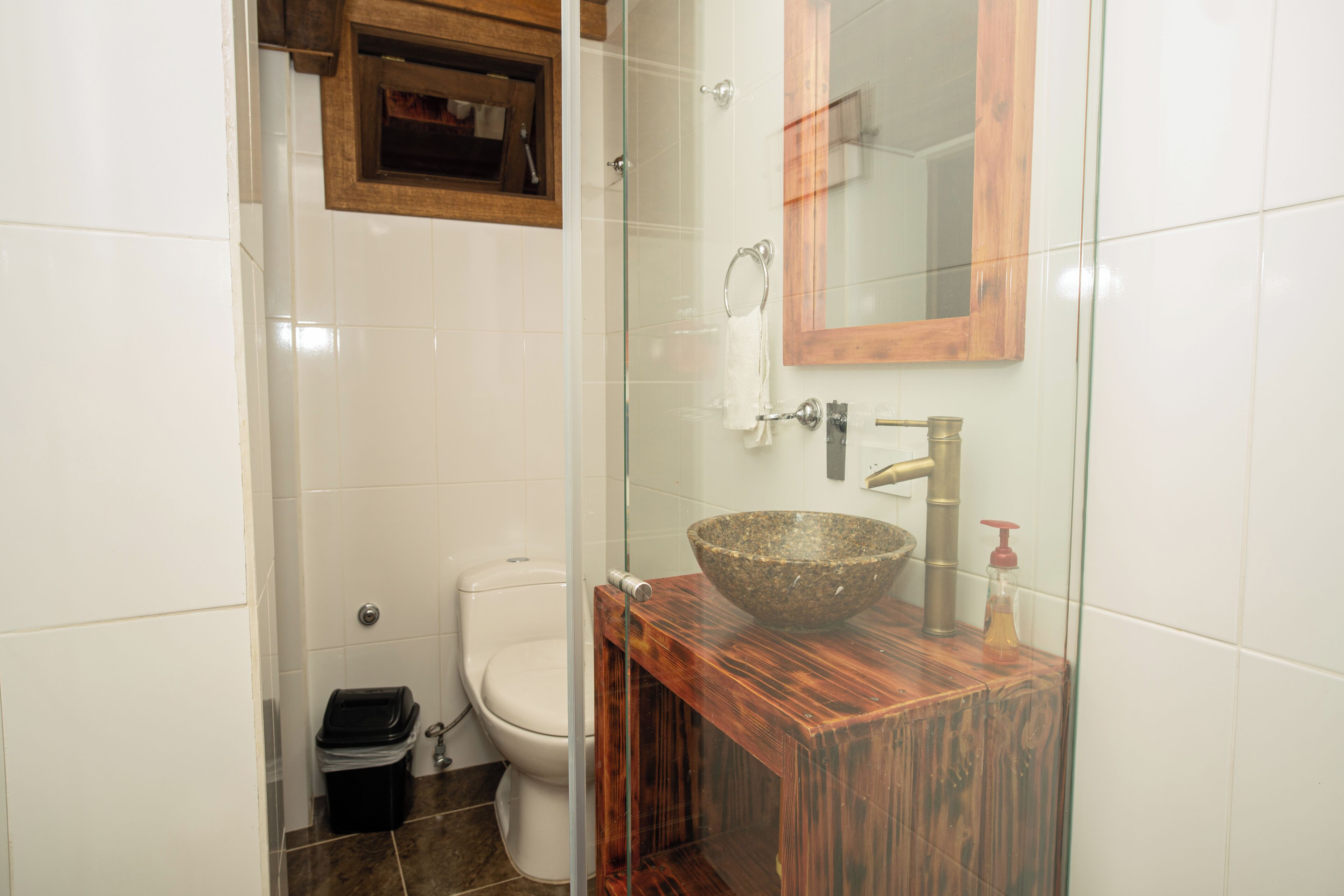 Et badeværelse med brusekabine i glas, et træbord, et hvidt toilet og et spejl med træramme.