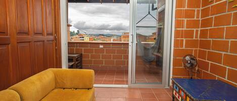Una habitación con un sofá amarillo, una pequeña mesa con bloques de colores, un ventanal corredero que da a un balcón y una pared de ladrillo.