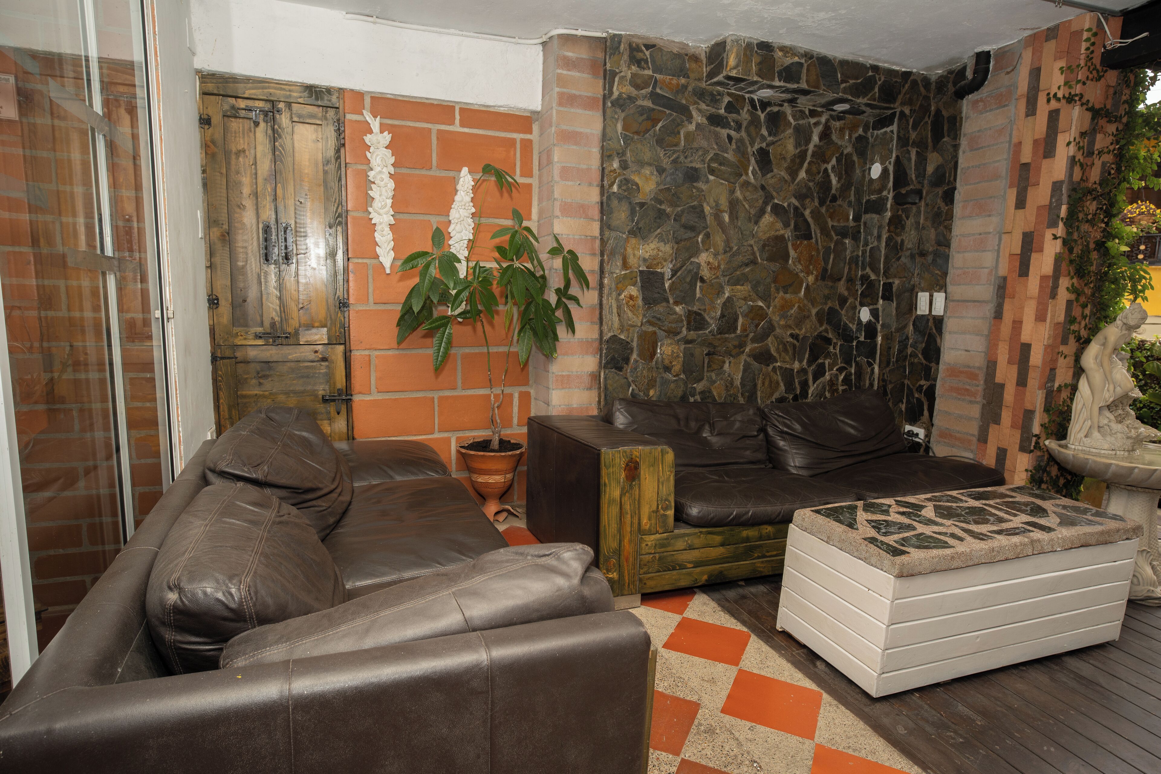 A living room with a brown leather sofa, a white ottoman, a potted plant, and a stone wall.