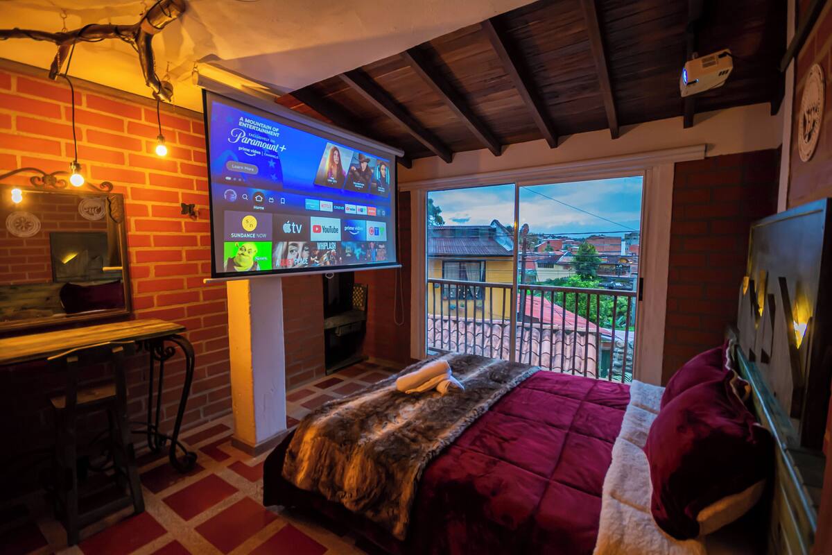 Quarto com parede de tijolos, uma grande tela de TV exibindo a interface de um serviço de streaming, uma varanda com vista para o exterior e uma cama com cobertor vermelho.