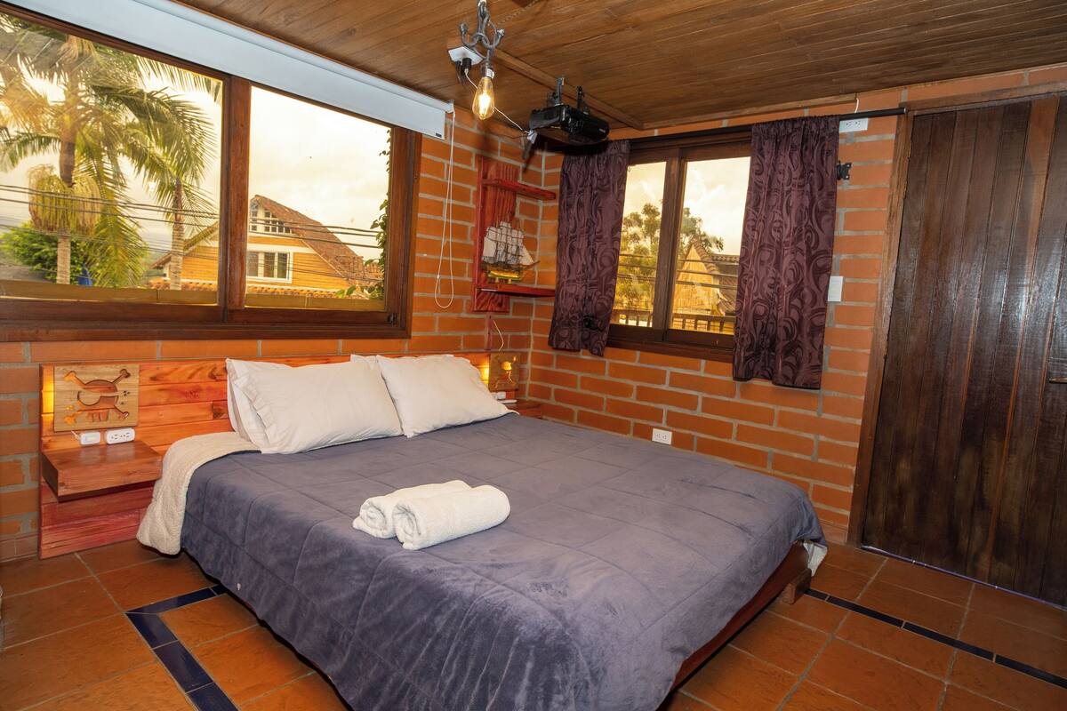 Quarto com cama grande, cabeceira de madeira, paredes de tijolo e janela com cortinas.