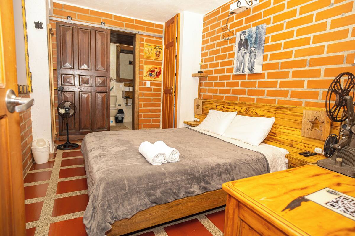 Quarto de hotel com cama, cabeceira de madeira, mesa de cabeceira, ventilador, parede de tijolos e porta que dá para outro quarto.