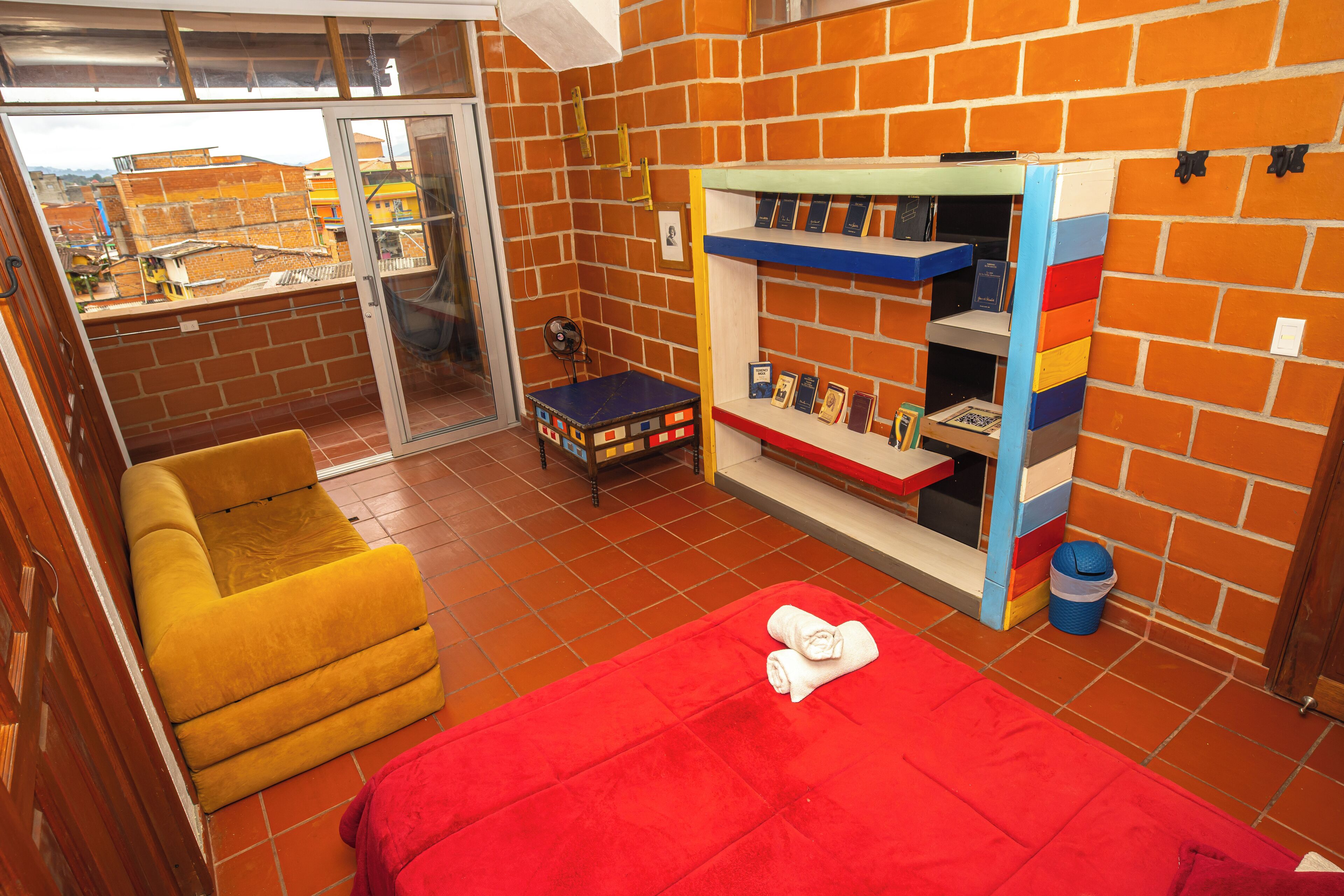 A room with a red bed, a yellow chair, a bookshelf with books, and a view through a glass door.