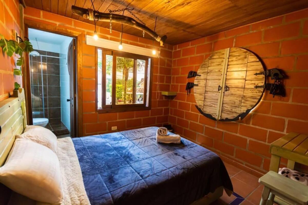 Quarto com cama, espelho redondo fixado na parede, teto de madeira e parede de tijolos.
