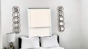 A hotel room with a bed, two white pillows, a bedside lamp, and a wall-mounted circular light fixture. - Renovated home 5 min downtown
 (San Antonio)