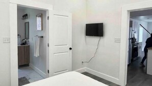 A modern, minimalistic room with a flat-screen TV mounted on the wall, a white door, and a visible bathroom area with a towel hanging.