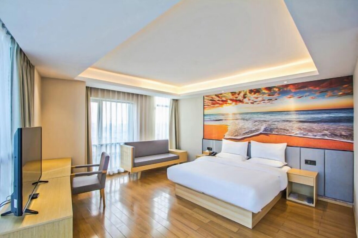 A modern hotel room with a large bed, a desk with a computer, a sofa, and a painting of a sunset over the ocean on the wall.