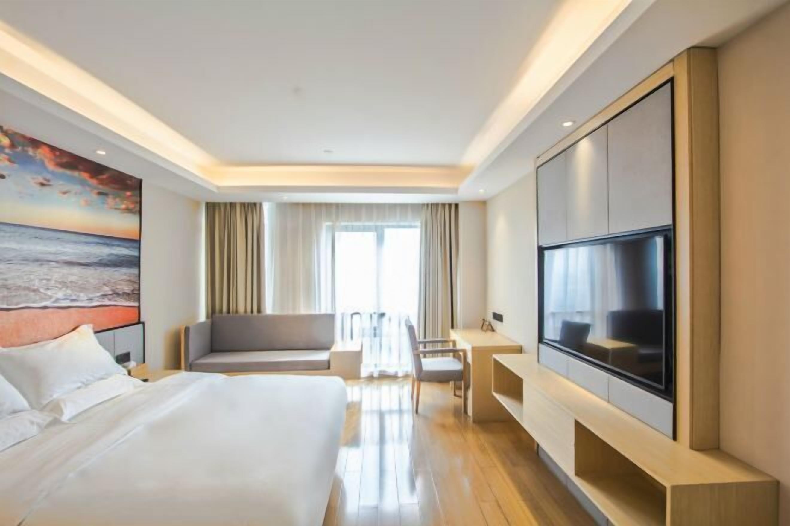 A modern hotel room with a large bed, a seating area, a desk, and a TV mounted on the wall.
