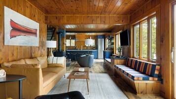 A cabin interior with a wooden ceiling, a sofa, a dining table, a kitchen area, and a large window.