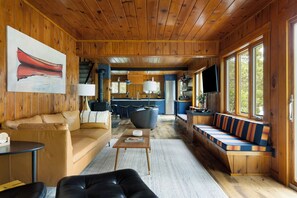 Ein Interieur einer Hütte mit Holzbalkendecke, einer Couch, einem Esstisch, einer Küchenzeile und einem großen Fenster.