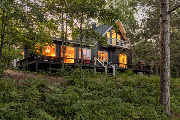 A cabin with a deck, large windows, and a staircase leading to a balcony.